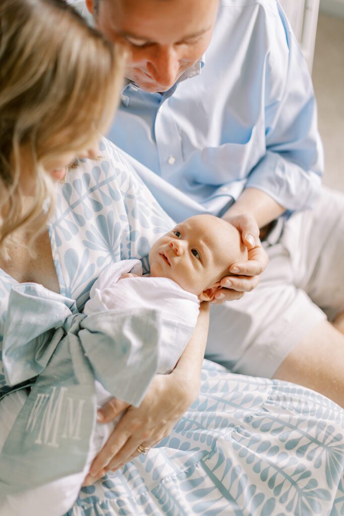 A mother holds her newborn baby wearing a swaddle with a monogrammed blue bow while dad gently places a hand on his head.