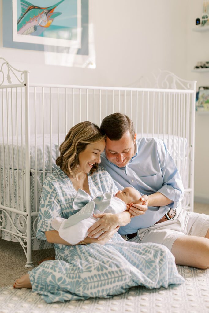 Parents holding their newborn baby sit on the floor next to a crib in a blue and white nursery.