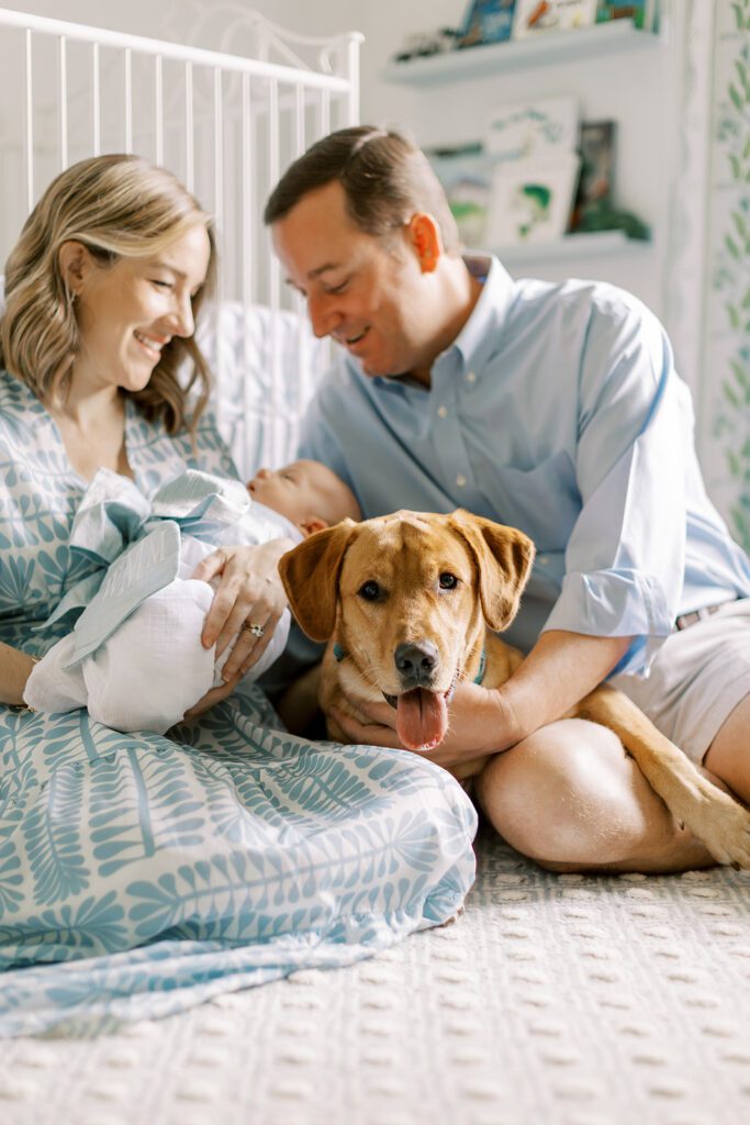 The parents a newborn baby are joined by their dog on the floor of a blue and white nursery during an at-home newborn photography session.