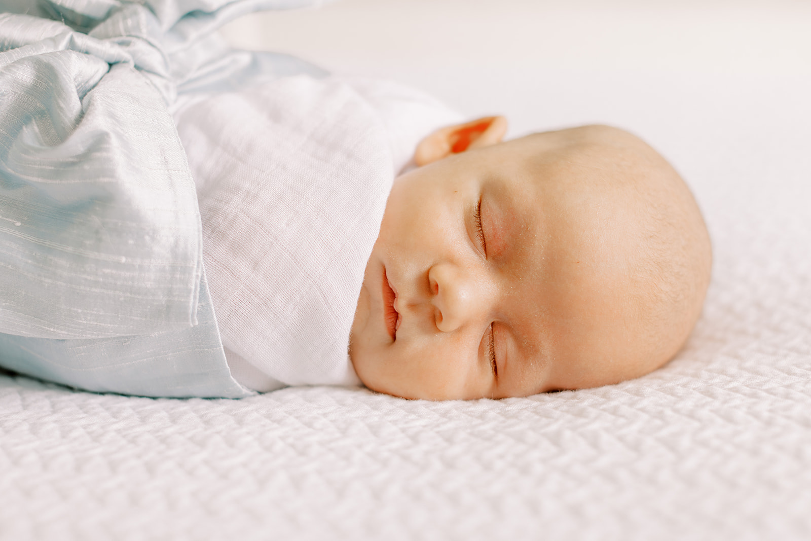 A newborn baby in a swaddle with a monogrammed blue blow sleeps on a white bed in natural light during a newborn photography sessinon.