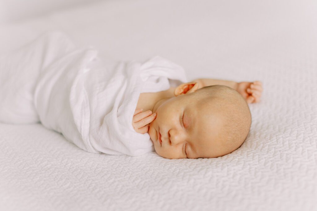 A newborn baby loosely wrapped in a white, cotton swaddle is sleeping on a white bed spread in natural light during a newborn photography session.